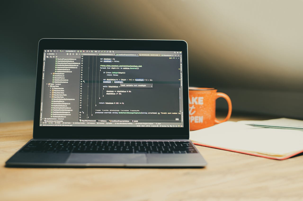 Laptop displaying coding software on a desk, alongside a coffee mug and notebook.